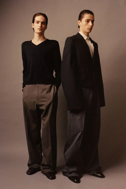 Two young adult men in dark casual and formal fashion standing in front of a neutral studio backdrop in a fashion editorial style.