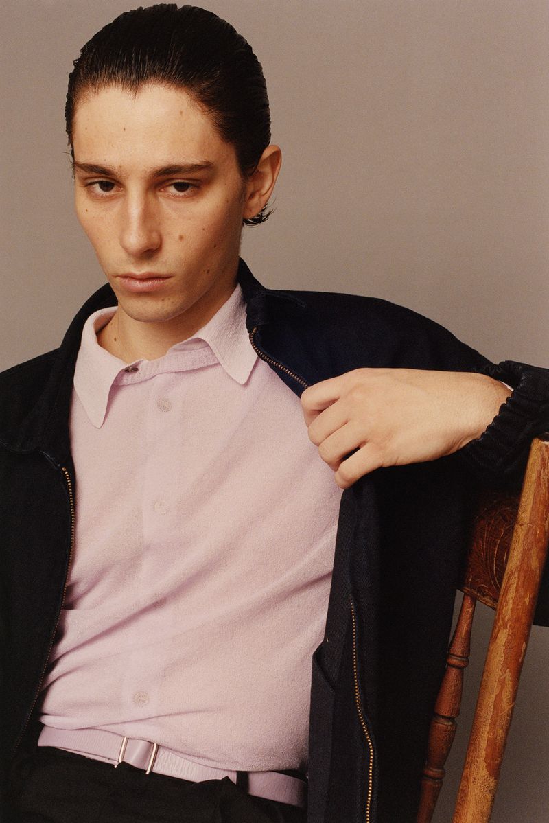 Portrait of a young adult man wearing a lavender shirt and navy jacket, sitting on a wooden chair against a plain gray background.