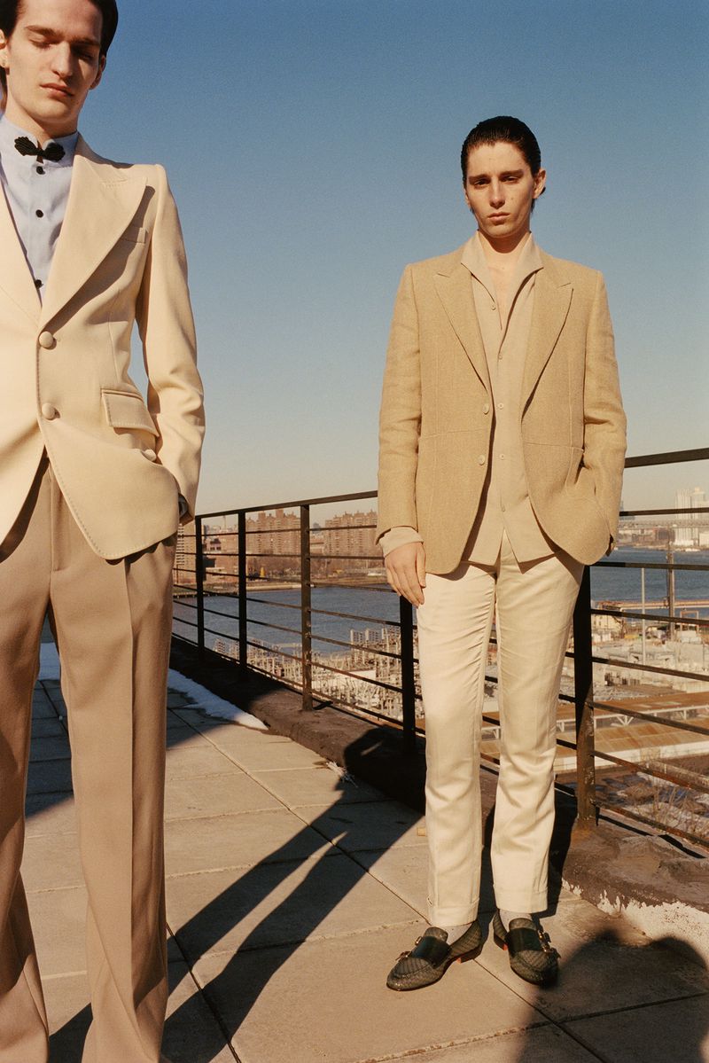 Two adult males in tan suits pose outdoors on a rooftop with city skyline and water in the background on a sunny day.
