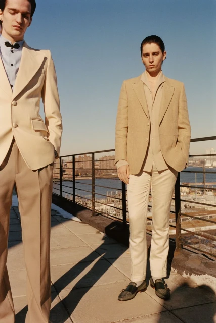Two adult males in tan suits pose outdoors on a rooftop with city skyline and water in the background on a sunny day.