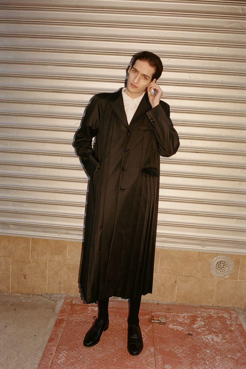 An adult male poses in a long black coat, black pants, and black shoes in front of a gray metal wall outdoors with a tiled floor.
