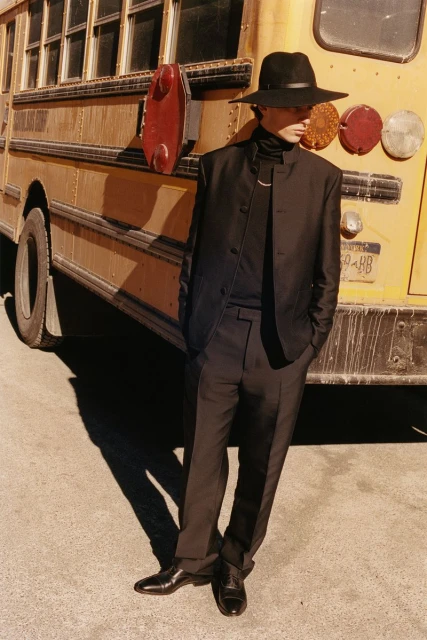 An adult male in stylish dark clothing poses next to a yellow school bus on pavement, bathed in warm sunlight, creating a striking contrast.