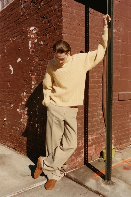 Male fashion model in a yellow sweater and tan trousers leans against a brick wall in an urban setting, posing in sunlight.