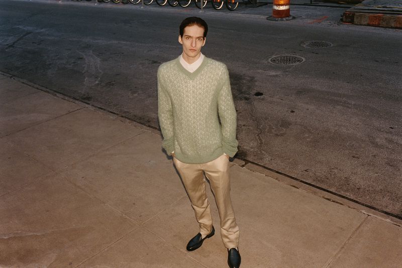Adult man stands on city sidewalk wearing a green patterned sweater, beige trousers, and black loafers in an eye-level, outdoor shot.