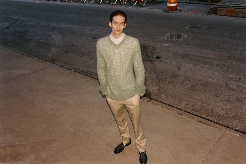 Adult man stands on city sidewalk wearing a green patterned sweater, beige trousers, and black loafers in an eye-level, outdoor shot.