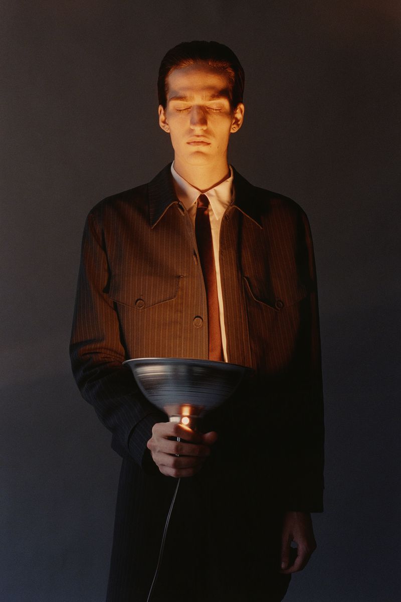 Adult male with closed eyes holds an illuminated gray bowl while wearing a striped jacket, white collared shirt, and brown tie in a studio.