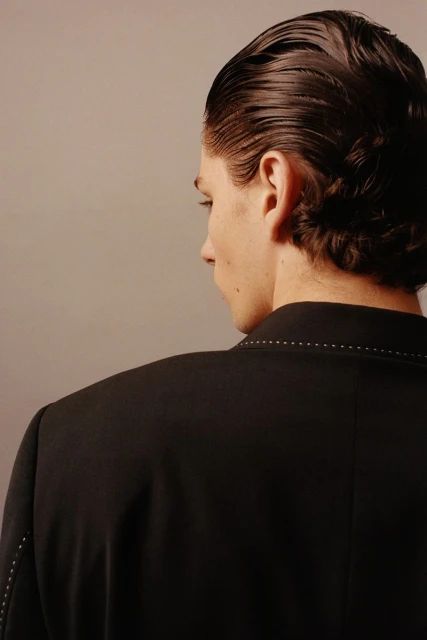 Man in a black suit poses in front of a neutral background, showcasing a slicked-back hairstyle and tailored fashion details.
