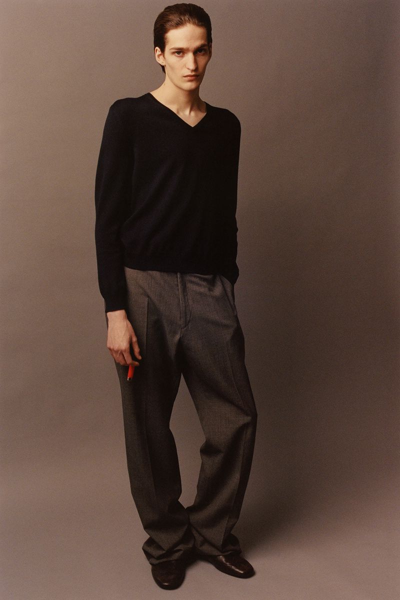 A slender man in a dark V-neck sweater and grey trousers stands on a solid grey background, holding a red object in his hand.