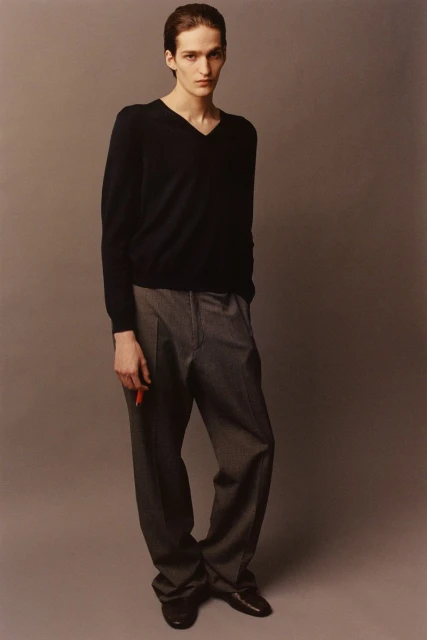 A slender man in a dark V-neck sweater and grey trousers stands on a solid grey background, holding a red object in his hand.
