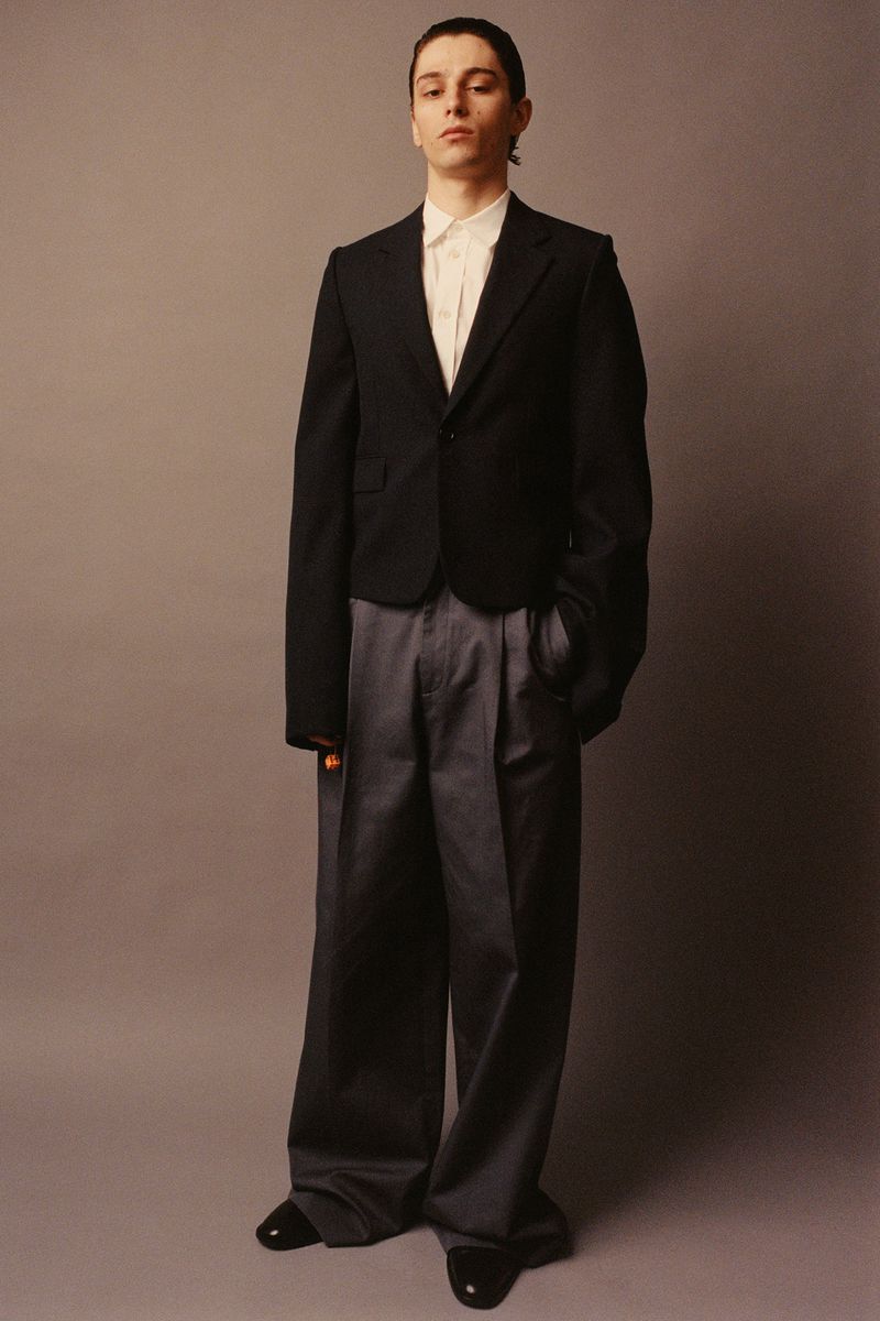 Full-length shot of a young adult man in a black blazer, white shirt, and gray slacks, standing against a solid neutral gray backdrop.