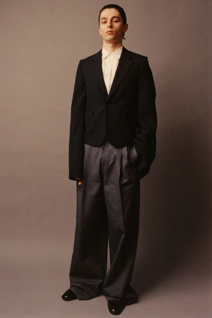 Full-length shot of a young adult man in a black blazer, white shirt, and gray slacks, standing against a solid neutral gray backdrop.