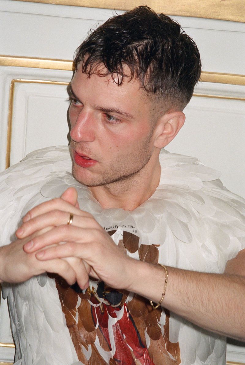 Close-up of a man wearing a feather-themed garment, a gold ring, and a gold bracelet, set against an indoor background with ornate molding.