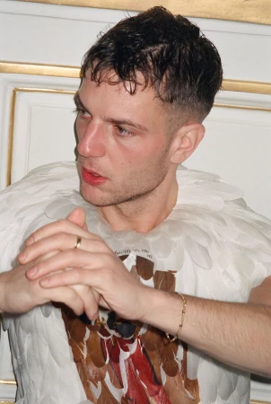 Close-up of a man wearing a feather-themed garment, a gold ring, and a gold bracelet, set against an indoor background with ornate molding.
