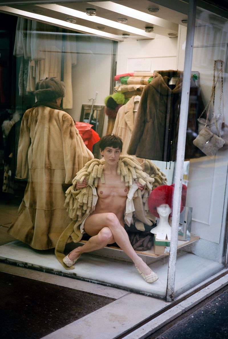 A nude woman wearing a coat is posed in a storefront window display with other coats and a mannequin in the background.