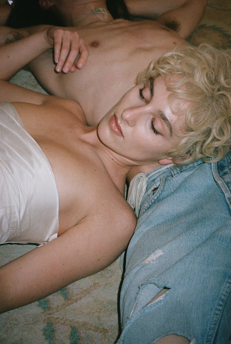 Intimate portrait of a woman with curly blonde hair resting on a person's lap while lying on a patterned carpet with soft, warm lighting.