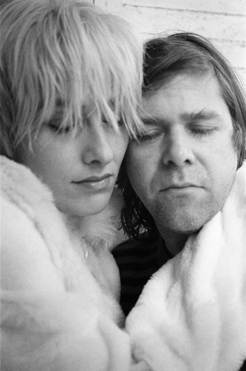 Black and white portrait of a serene couple embracing and wearing matching fur coats, their eyes gently closed in a moment of calm.