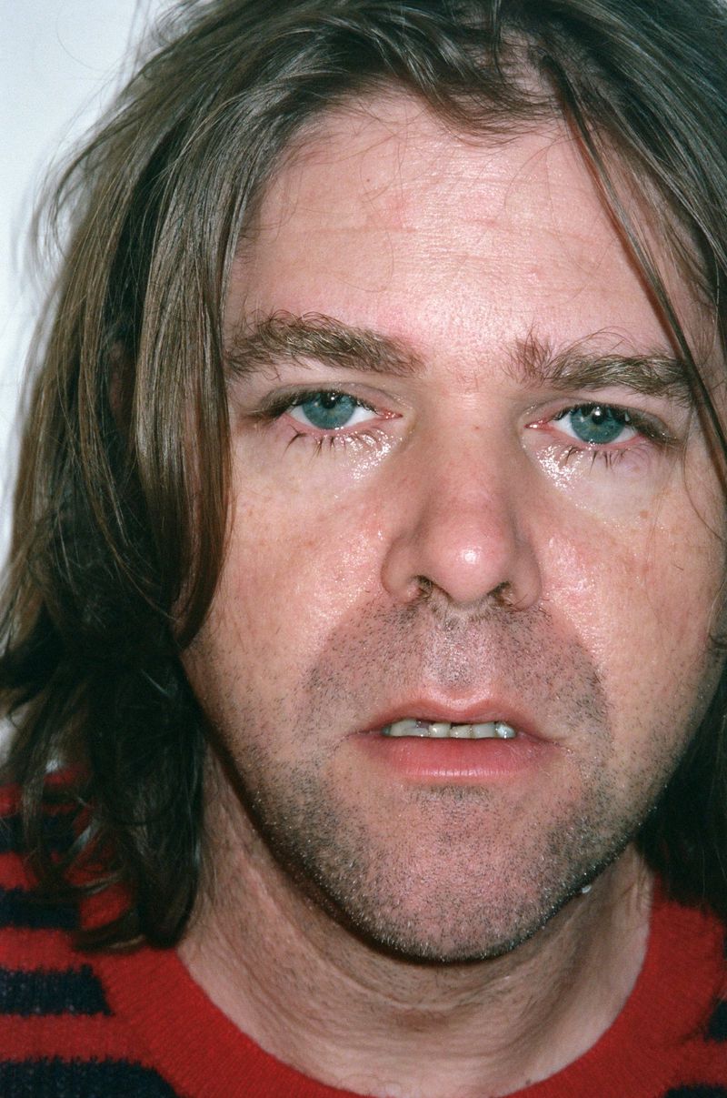 Close-up portrait of a man with shoulder-length brown hair, light stubble, and blue eyes, wearing a red and black striped sweater.