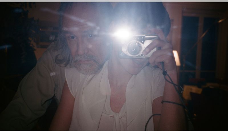 Selfie of two people indoors. One adult male with grey hair is partially visible, holding a camera with flash, while the second person is behind.