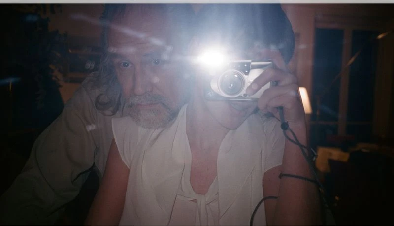 Selfie of two people indoors. One adult male with grey hair is partially visible, holding a camera with flash, while the second person is behind.