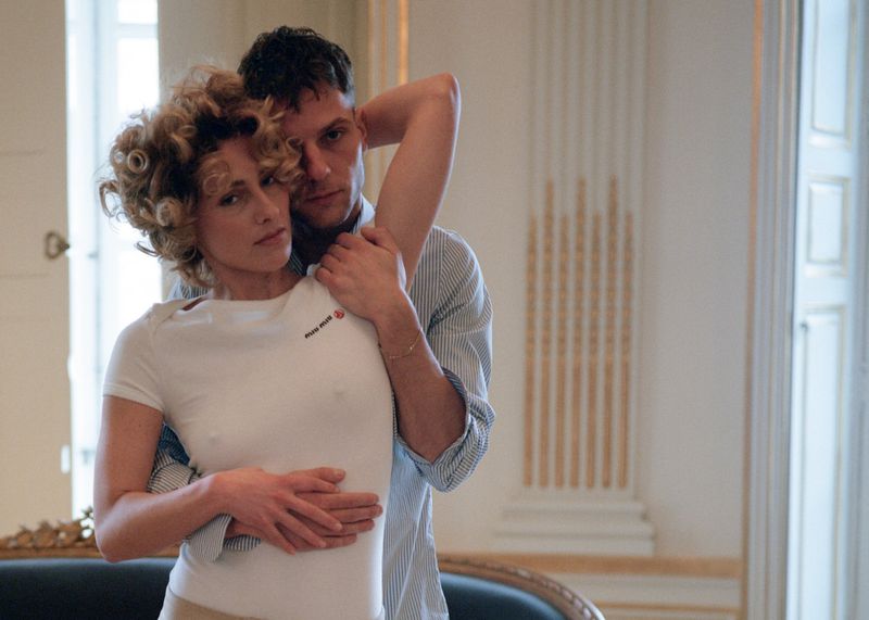 A woman with curly blonde hair is embraced from behind by a man wearing a striped shirt, in an indoor setting with natural light.