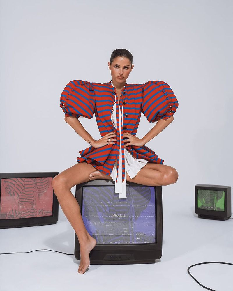An adult woman sits cross-legged atop a black television with additional televisions in a bright photo studio environment.