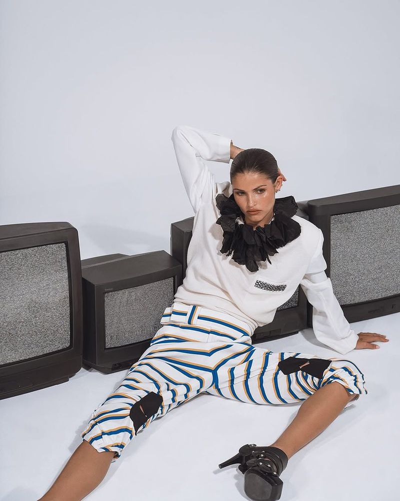 A fashionable adult female sits on the floor next to static-filled retro TVs, wearing a white sweater and striped pants in a studio setting.