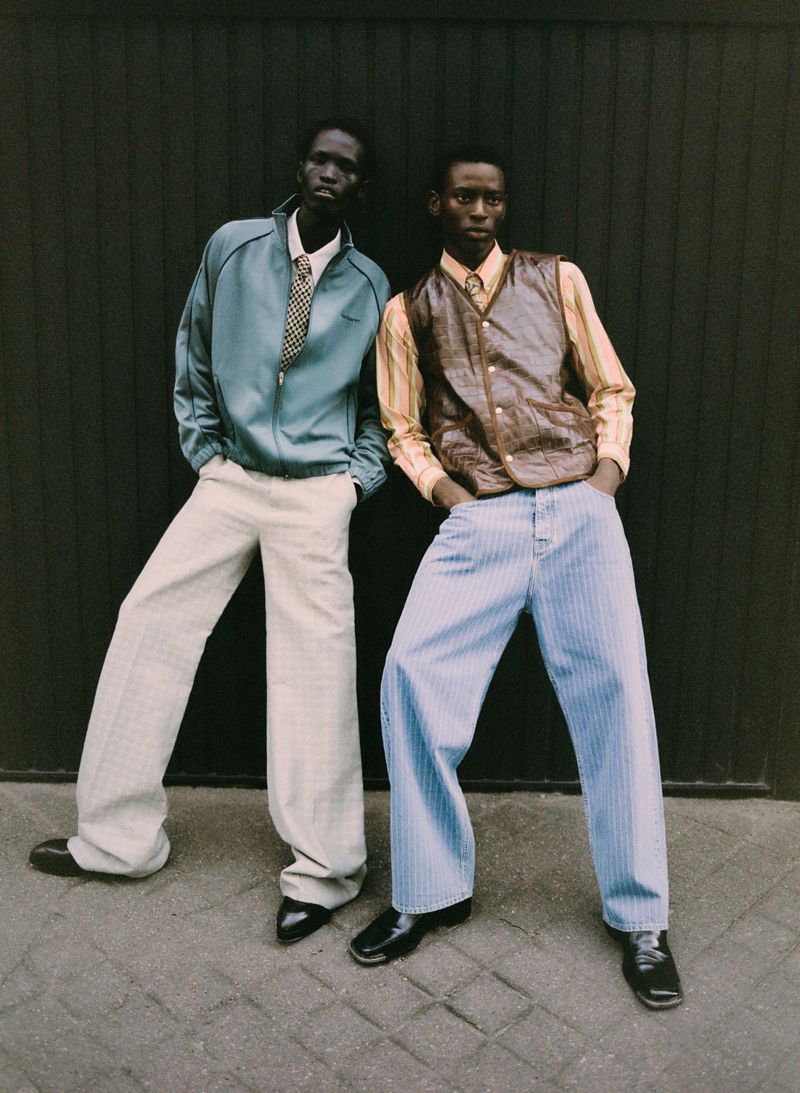 Two black men in fashionable vintage-inspired attire pose against a dark gray wall, showcasing pale gray wide-leg pants and striped pants.