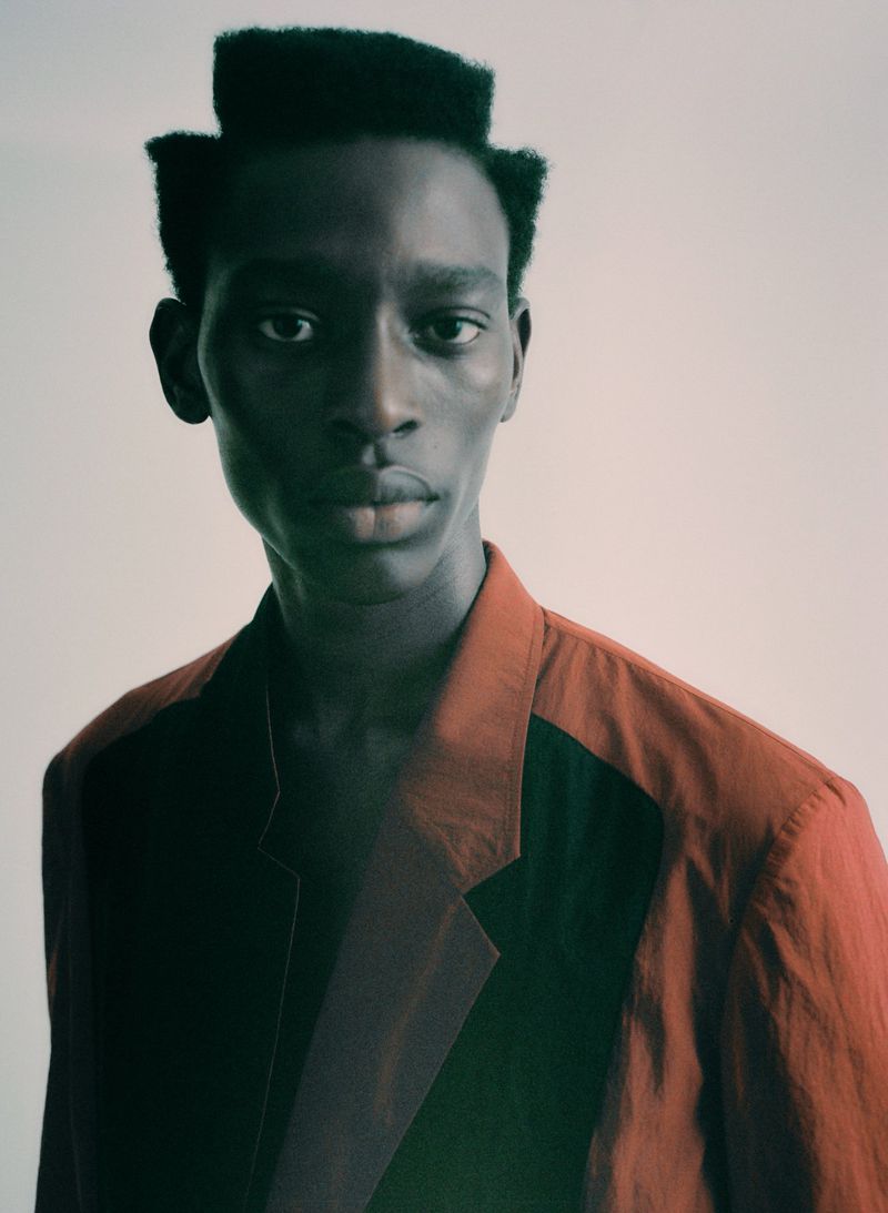 Portrait of a Black male model with a unique haircut wearing a rust-colored blazer against an off-white studio backdrop, fashion-forward aesthetic.
