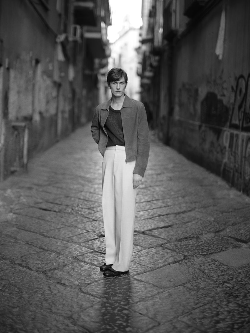 A young adult man stands in a narrow cobblestone street, wearing a jacket and white pants, in a black and white fashion shot.