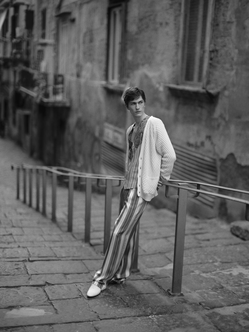 Male model in striped pajamas and a cardigan poses on stone steps with a metal railing in an outdoor urban setting in black and white.