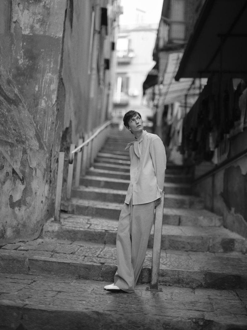 Black and white portrait of a young adult woman in a stylish outfit looking up while posing on stone steps in a narrow urban alley.