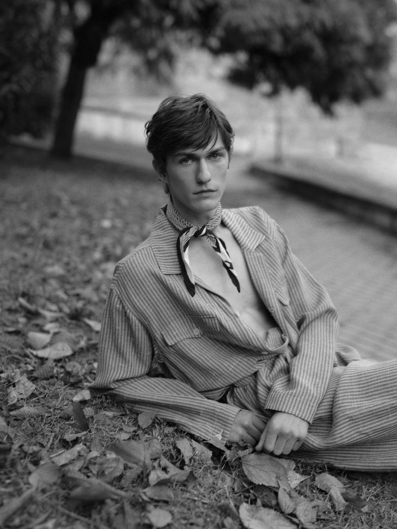 Monochrome portrait of a young adult male model lying on the ground in a striped suit and a patterned scarf, posing in an outdoor setting.