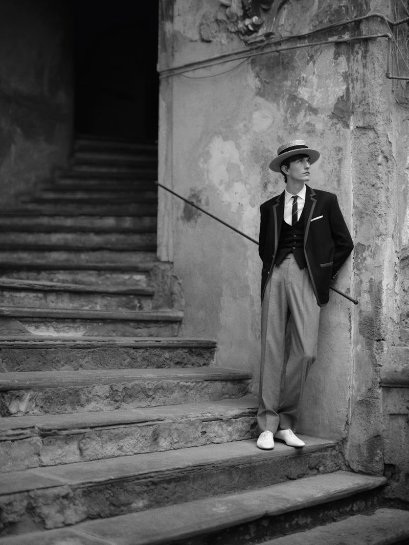 Black and white portrait of a stylish young adult male leaning against a weathered stone stairway outdoors, wearing a suit and boater hat.