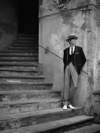 Black and white portrait of a stylish young adult male leaning against a weathered stone stairway outdoors, wearing a suit and boater hat.