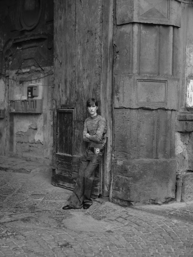 Young man in patterned sweater poses against weathered stone wall and arched doorway in black and white, creating a vintage street style.