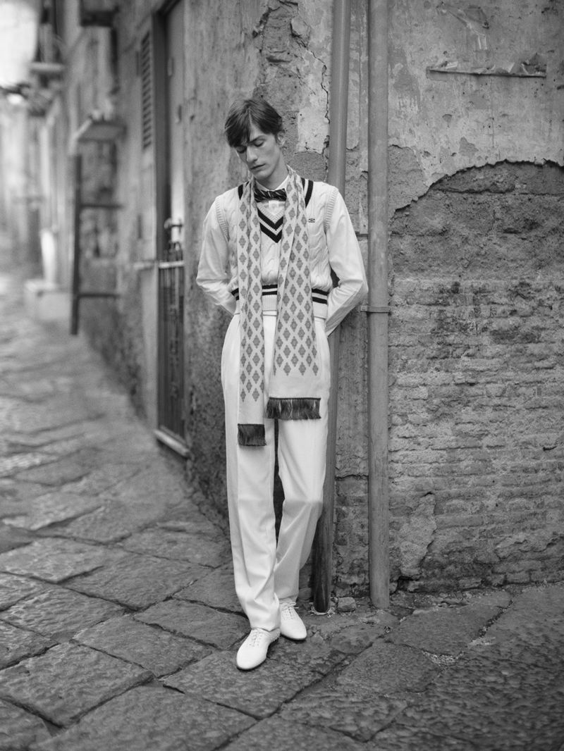 Black and white portrait of a young adult man leaning against a weathered wall in a narrow outdoor urban setting, wearing a scarf and suit.