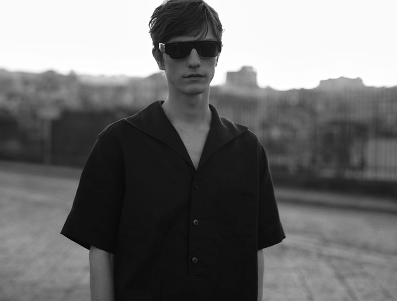Monochrome portrait of a young man wearing sunglasses and a dark button-down shirt outdoors against a blurred cityscape.