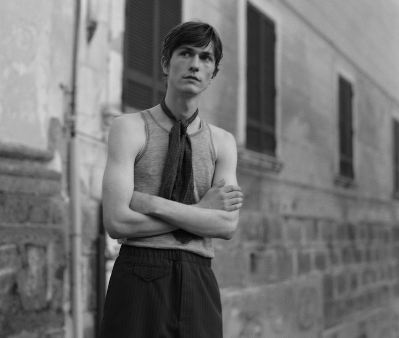 Black and white portrait of a male model with crossed arms, wearing a tank top and scarf, against a building backdrop.