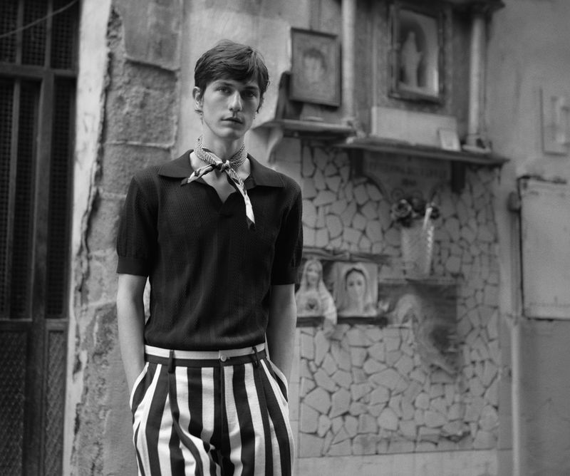 Monochrome portrait of a young adult male wearing a neck scarf, posing outdoors against an old wall with decorative elements.