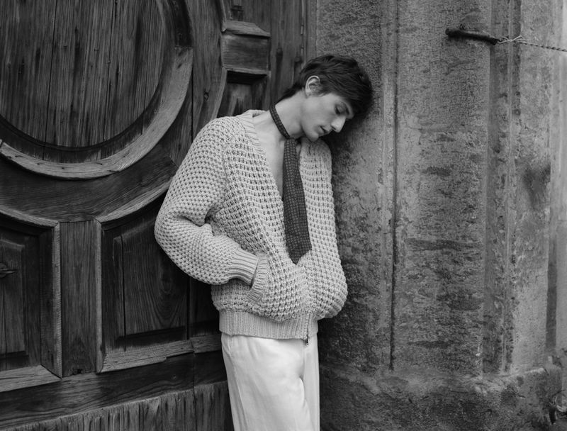 Monochrome portrait of a young man leaning against a weathered stone wall beside a vintage wooden door, wearing a knit cardigan and tie.