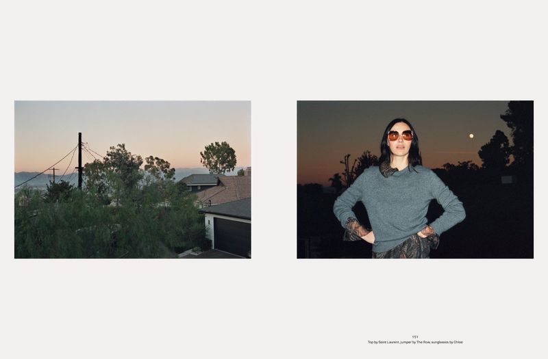 Fashion editorial shot of a woman wearing sunglasses and a grey sweater, standing outdoors at sunset. Landscape view of homes.