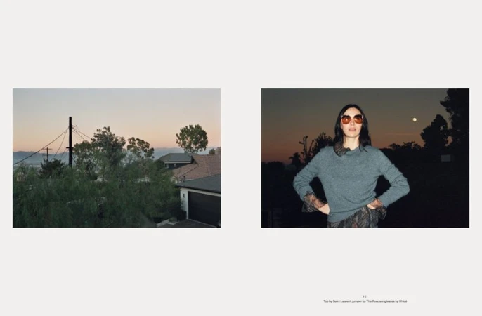 Fashion editorial shot of a woman wearing sunglasses and a grey sweater, standing outdoors at sunset. Landscape view of homes.