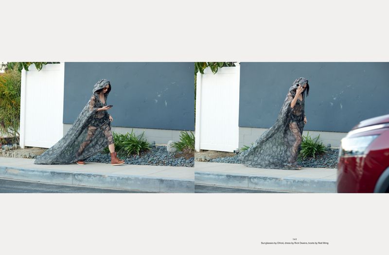 Fashion shot of a woman in sunglasses wearing a hooded lace dress and boots walking on a sidewalk next to a building with greenery.