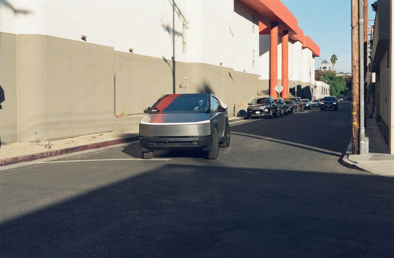A Tesla Cybertruck drives along a city street with buildings in the background on a sunny day, showcasing its unique design.