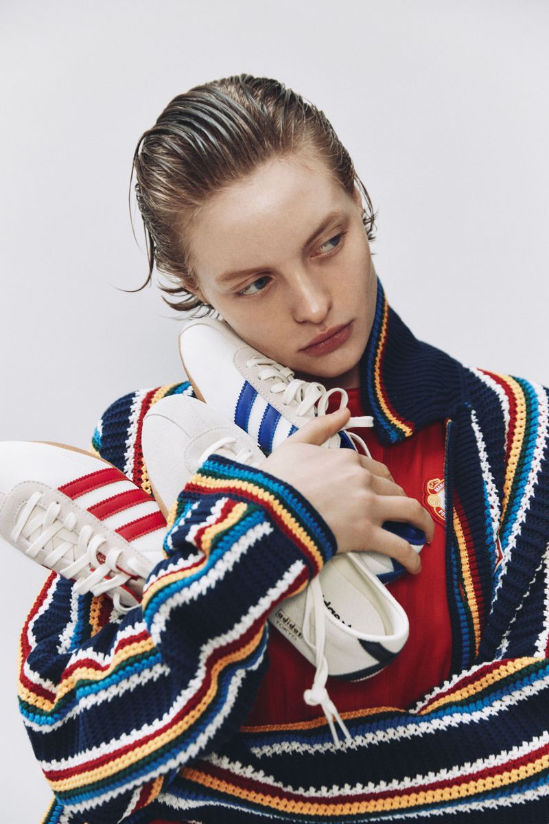 Model with slicked-back hair holding three pairs of sneakers against a plain white background for LuisaViaRoma and Adidas.