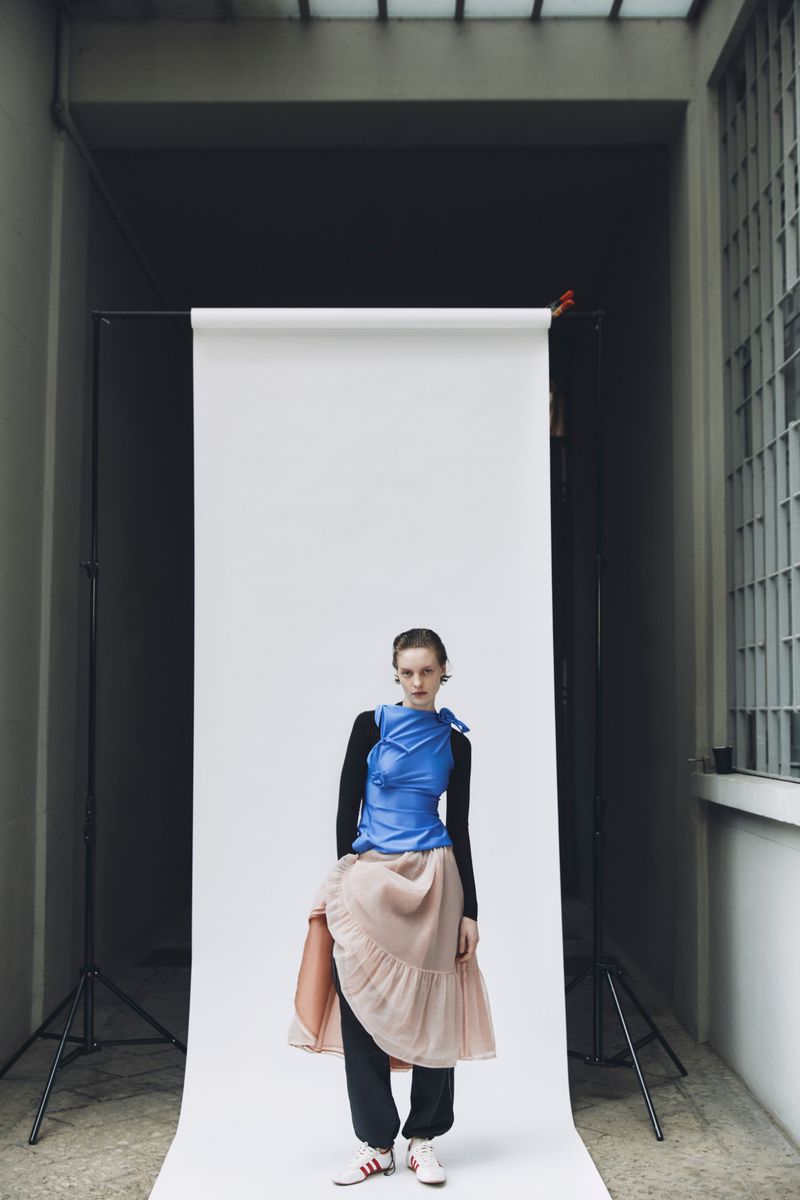 A young adult woman poses on a white backdrop wearing a blue top, long sleeve shirt, skirt, sweatpants, and sneakers in an indoor setting.