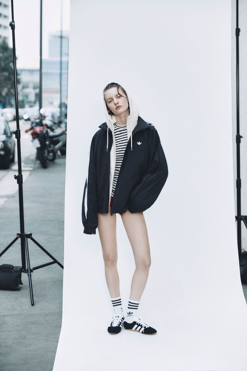 Fashion photography of a female model wearing an Adidas jacket, striped top, socks, and sneakers against a white background indoors.