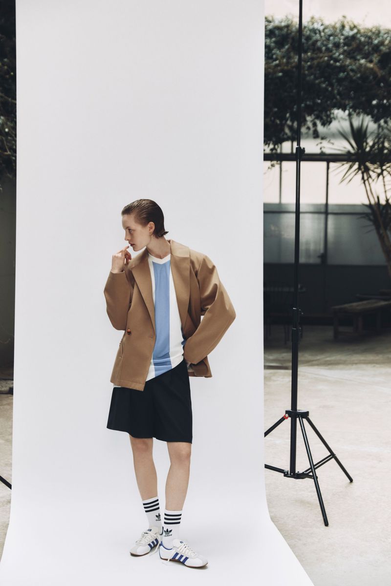 A young adult in a tan blazer, blue and white striped shirt, and black shorts stands in front of a white backdrop in a studio setting.