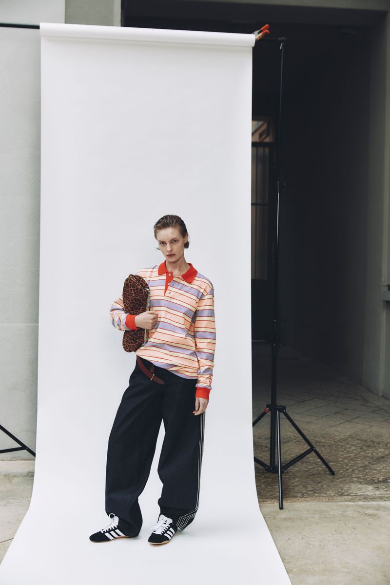 Woman in striped shirt and wide-leg pants poses in a studio with a brown patterned bag. Adidas sneakers and a white backdrop complete the scene.