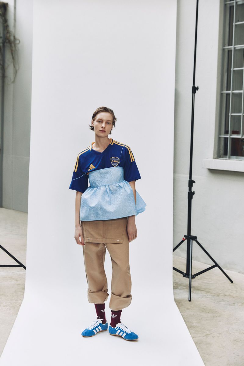 Fashion shot of a female model in an Adidas soccer jersey, striped top, beige pants, and blue shoes posing in front of a white wall.